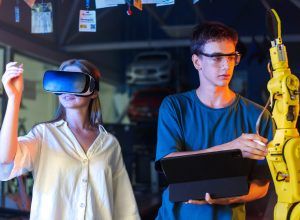 Teens doing experiments in robotics in a laboratory. Boy in protective glasses using tablet and girl in VR headset controlling the robot using her hand. Red and blue illumination
